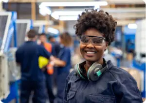 African American woman smiling in an industrial setting, wearing safety goggles and headphones.